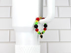 A beaded bracelet with red, yellow, green, and black beads. The bracelet has a gold-colored heart charm. The bracelet is displayed on a white mug handle against a white tile background.