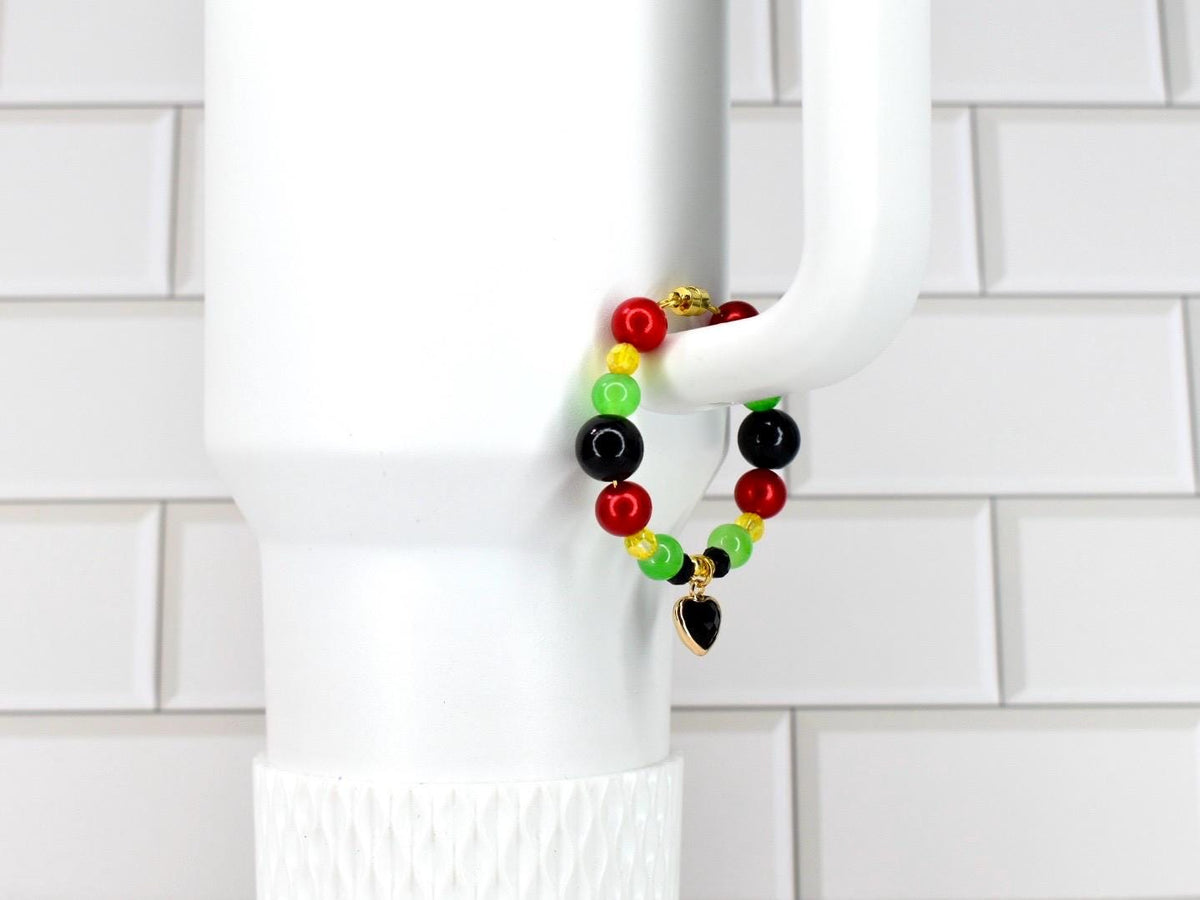 A beaded bracelet with red, yellow, green, and black beads. The bracelet has a gold-colored heart charm. The bracelet is displayed on a white mug handle against a white tile background.