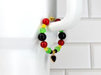 A beaded bracelet with red, yellow, green, and black beads. A gold-colored heart charm with a black center hangs from the bracelet. The bracelet is displayed against a white background.