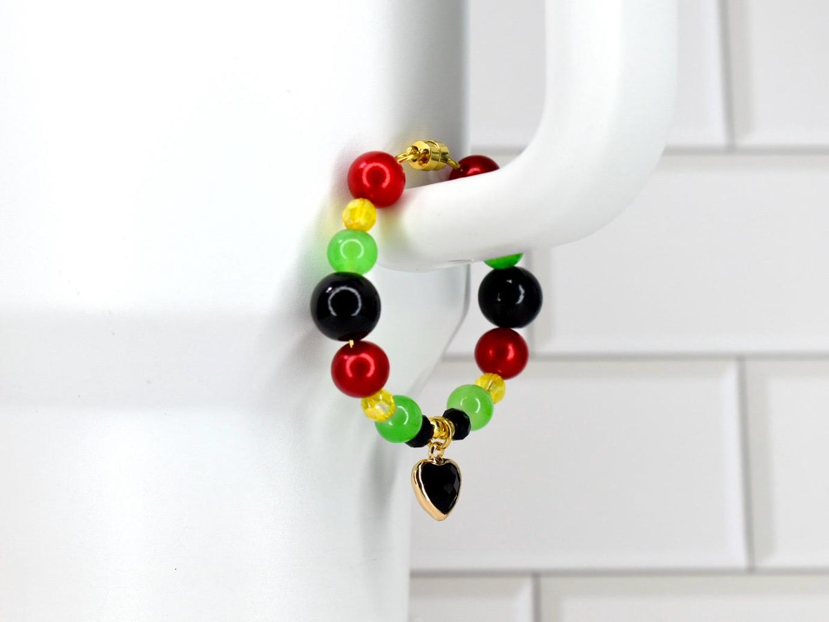 A beaded bracelet with red, yellow, green, and black beads. A gold-colored heart charm with a black center hangs from the bracelet. The bracelet is displayed against a white background.