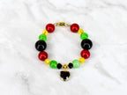 A beaded bracelet featuring red, yellow, green, and black beads. The bracelet has a gold-colored magnetic clasp and a heart-shaped charm with a black center. The beads are round and evenly spaced.