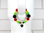 A beaded bracelet featuring red, green, black, and yellow beads. The bracelet has a gold-colored heart charm with a black center. The beads are strung together, creating a colorful accessory.