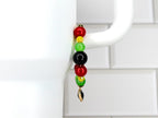A beaded tumbler handle bracelet with red, yellow, green, and black beads, and a gold-colored charm. The bracelet is hanging from the handle of a white mug. The beads are round and shiny.