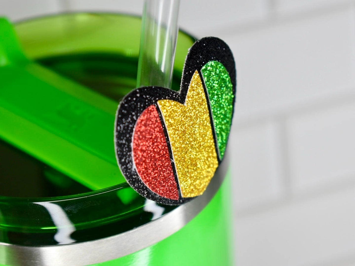 A heart-shaped glitter charm in red, yellow, green, and black accents, attached to a green tumbler with a straw. The charm is a decorative accessory for drinkware, adding a touch of sparkle.