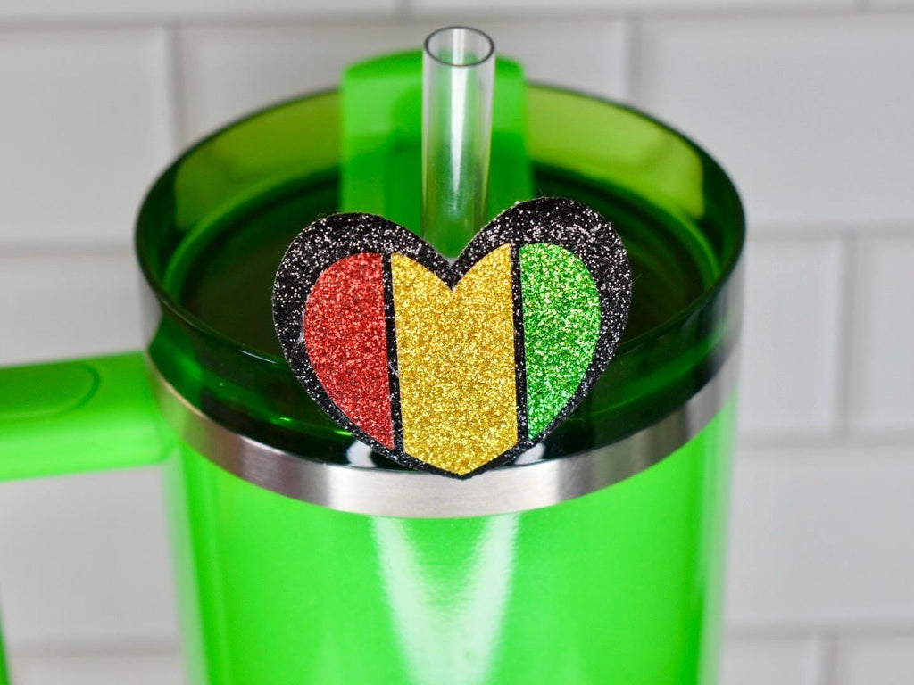 A green tumbler with a heart-shaped charm. The charm is divided into red, gold, and green glitter sections, outlined in black glitter. A clear straw is inserted into the lid. The tumbler has a handle.