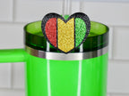 A green tumbler with a silver rim and handle, topped with a heart-shaped glitter charm. The charm features red, gold, and green vertical stripes, outlined in black, and is attached to a clear straw.