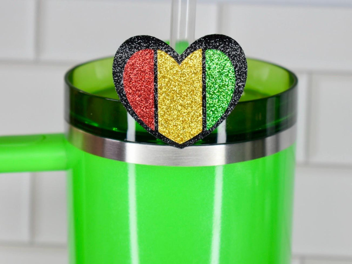 A green tumbler with a silver rim and handle, topped with a heart-shaped glitter charm. The charm features red, gold, and green vertical stripes, outlined in black, and is attached to a clear straw.