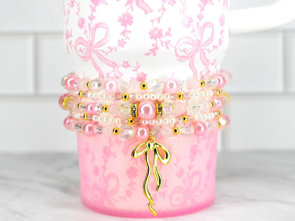 A pink and white floral-patterned cup with a stack of beaded bracelets. The bracelets feature pink pearls, clear beads, and gold accents. A gold bow charm hangs from the bracelets, adding a touch of elegance.