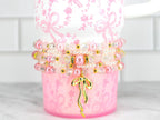 A pink and white floral-patterned cup with a stack of beaded bracelets. The bracelets feature pink pearls, clear beads, and gold accents. A gold bow charm hangs from the bracelets, adding a touch of elegance.