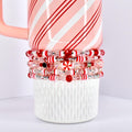 A stack of beaded bracelets in red, white, and clear colors. The bracelets are adorned with various beads, including candy cane-striped and clear beads. The bracelets are resting on a white ceramic cup with a pink and white striped mug.