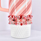 A stack of beaded bracelets in red, white, and clear colors. The bracelets are adorned with various beads, including candy cane-striped and clear beads. The bracelets are resting on a white ceramic cup with a pink and white striped mug.