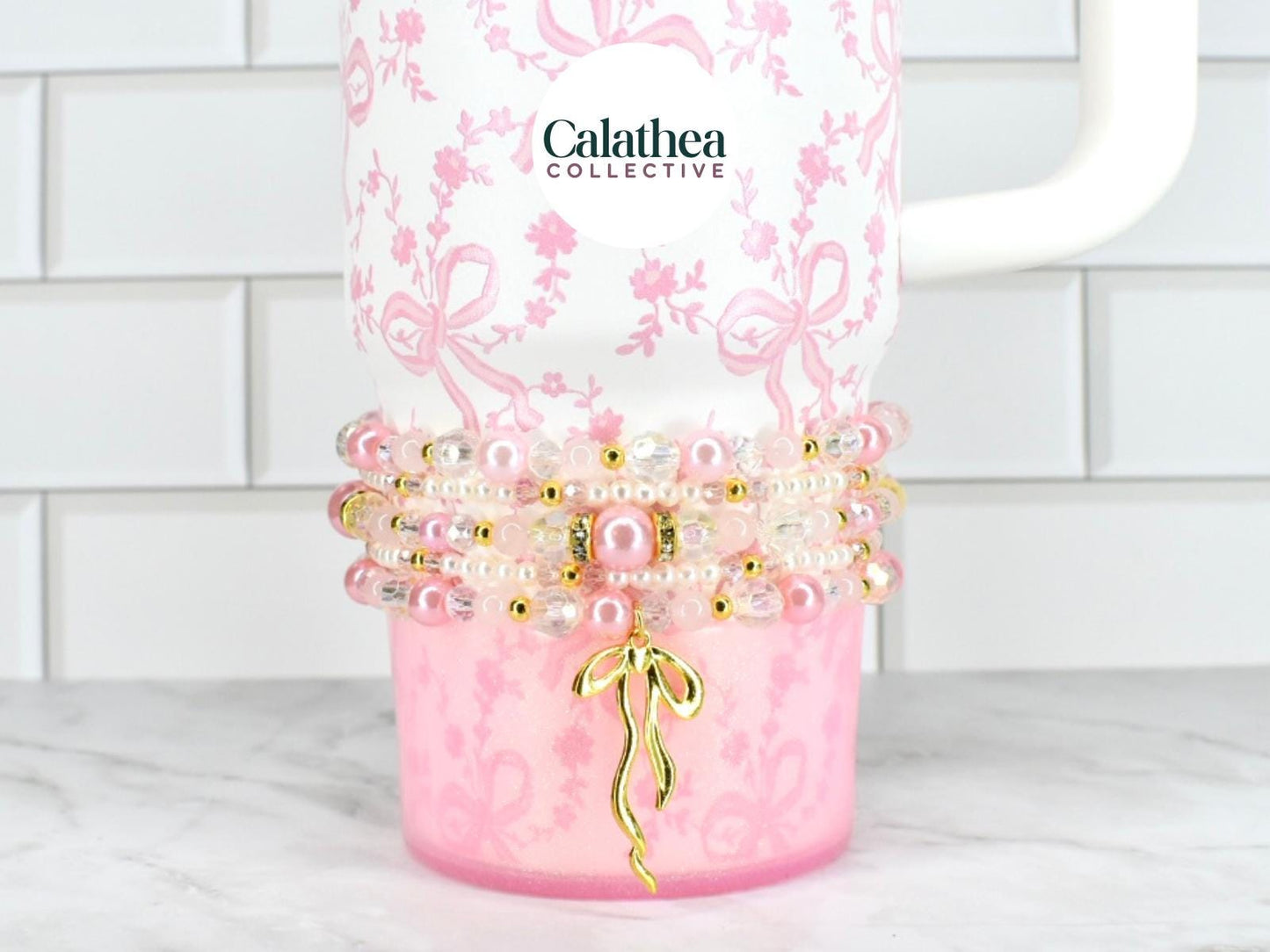 A white tumbler with a pink floral and bow pattern, adorned with beaded bracelets in shades of pink, pearl, and gold. A gold bow charm hangs from the bracelets. The brand name "Calathea Collective" is visible.