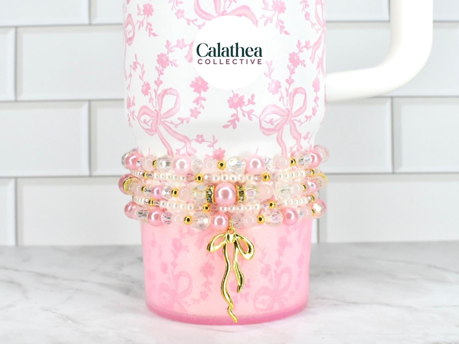 A white tumbler with a pink floral and bow pattern, adorned with beaded bracelets in shades of pink, pearl, and gold. A gold bow charm hangs from the bracelets. The brand name "Calathea Collective" is visible.