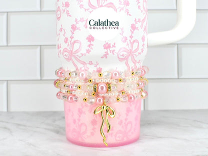 A white tumbler with a pink floral and bow pattern, adorned with beaded bracelets in shades of pink, pearl, and gold. A gold bow charm hangs from the bracelets. The brand name "Calathea Collective" is visible.