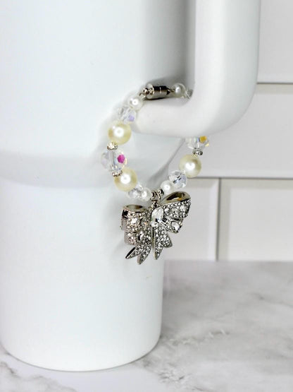 A white beaded bracelet with a silver bow charm. The bracelet features faux pearls and clear crystal beads. The bow charm is encrusted with small, sparkling crystals. The bracelet is attached to a white mug.