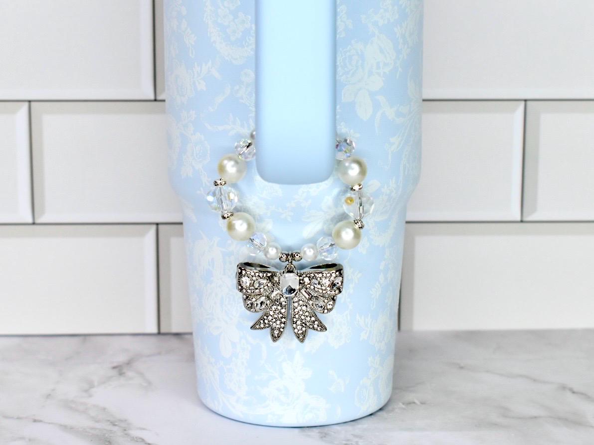 A light blue Stanley with a floral pattern and a decorative bow. The bow is silver with clear rhinestones. The tumbler has a pearl and crystal beaded strap.