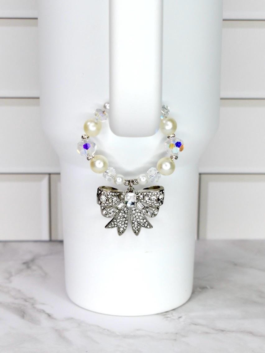 A decorative cup accessory featuring a silver bow adorned with clear rhinestones. The bow is attached to a beaded band with faux pearls and iridescent crystals. The accessory is designed to fit around a white cup.