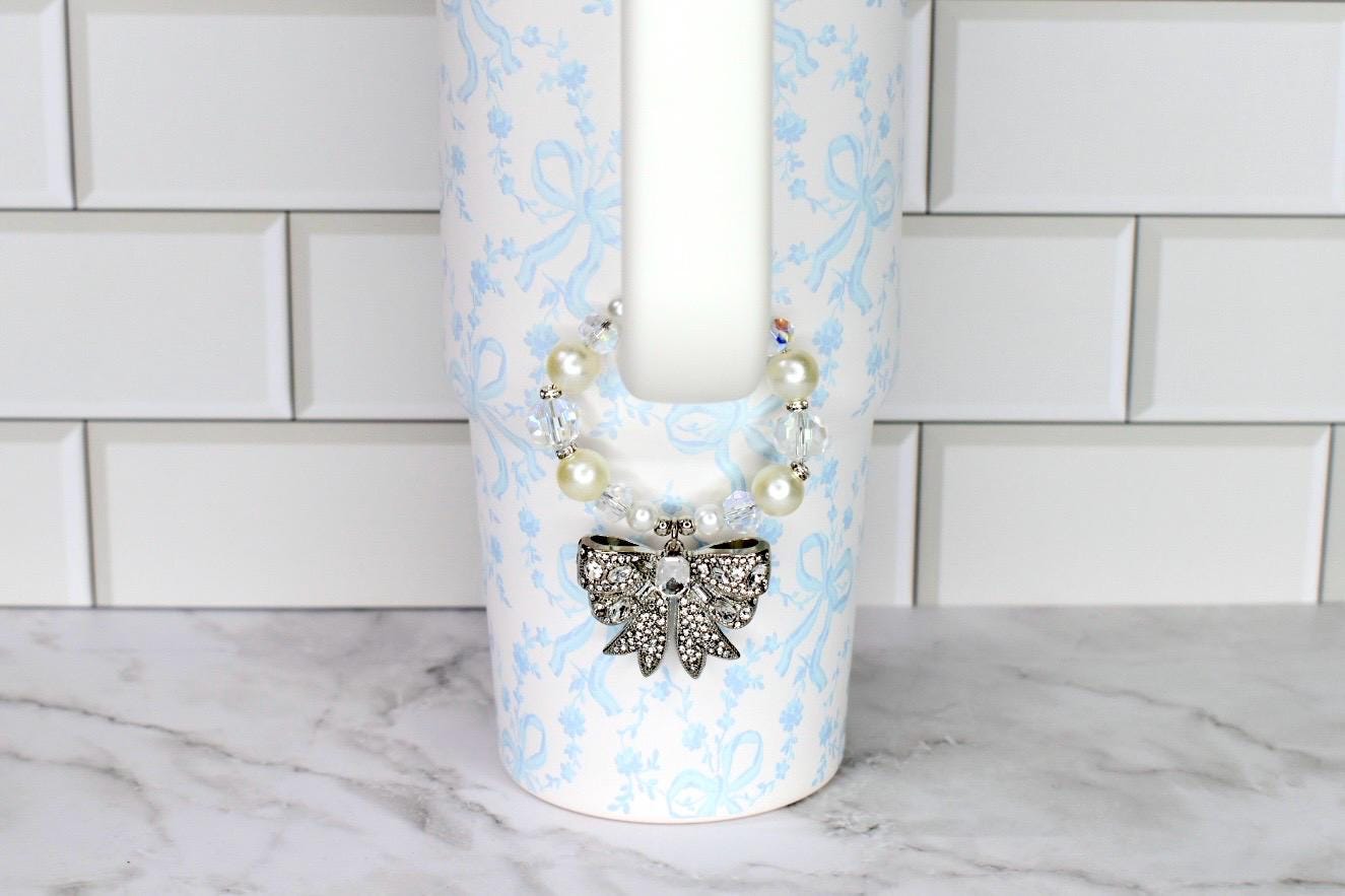 A white tumbler with a light blue floral and bow pattern. A beaded necklace with faux pearls and clear crystals is wrapped around the top. A silver bow pendant with clear rhinestones hangs from the necklace.