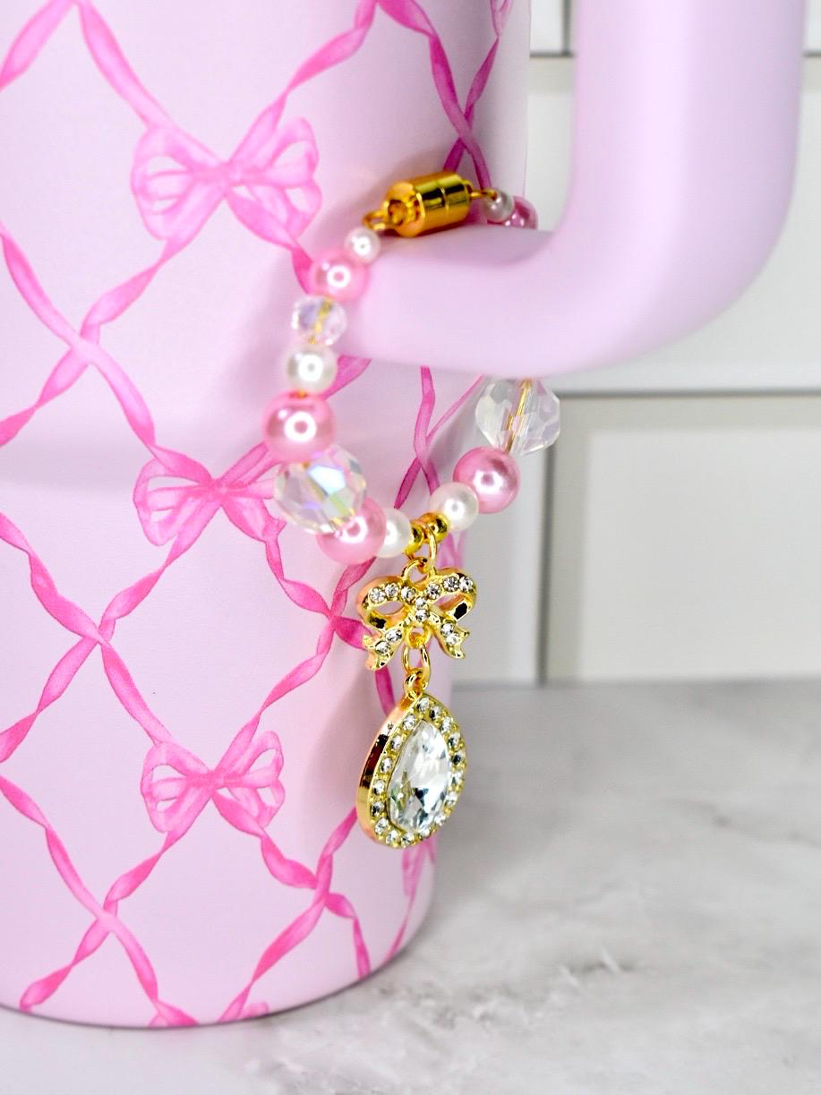 A pink beaded bracelet with a gold bow charm and a teardrop-shaped pendant with a clear stone. The bracelet is attached to a pink mug with a bow pattern. The bracelet features pink and clear beads.