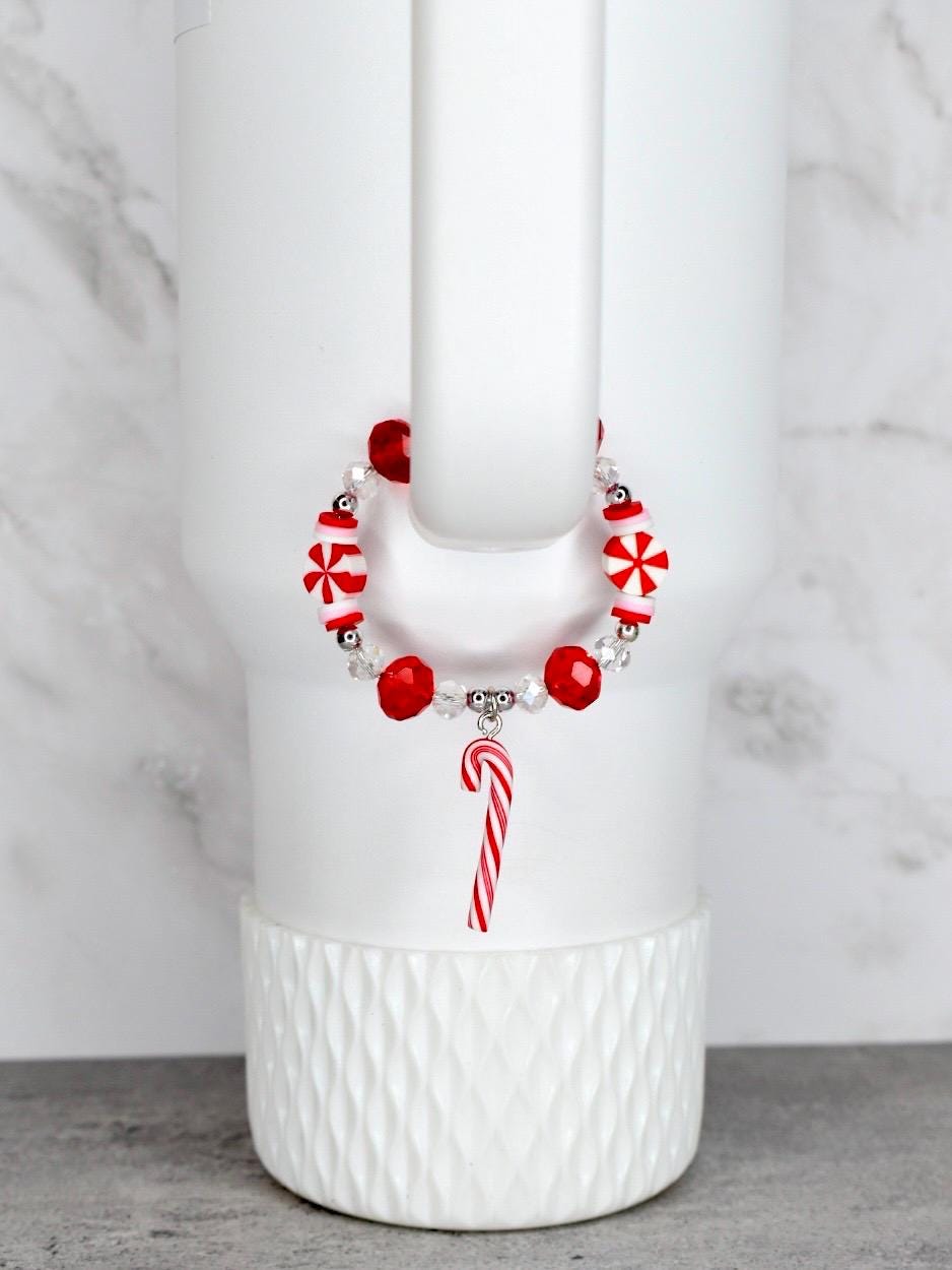 A festive bracelet with red and white candy-themed beads and a candy cane charm. The bracelet is strung on a clear elastic cord. The bracelet is displayed on a white tumbler.