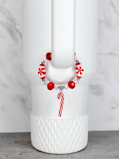 A festive bracelet with red and white candy-themed beads and a candy cane charm. The bracelet is strung on a clear elastic cord. The bracelet is displayed on a white tumbler.