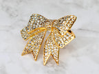 A gold-toned bow straw charm encrusted with numerous clear crystals. The bow design features detailed folds and pointed ends, creating a sparkling, elegant accessory. The charm is set against a marble-like background.