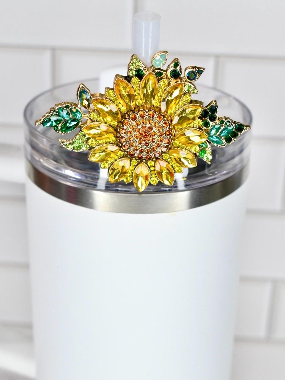 A white tumbler with a decorative sunflower lid. The sunflower is made of yellow and orange crystals with green crystal leaves. The tumbler has a clear lid and a white straw.