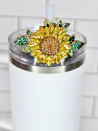 A white tumbler with a decorative sunflower lid. The sunflower is made of yellow and orange crystals with green crystal leaves. The tumbler has a clear lid and a white straw.
