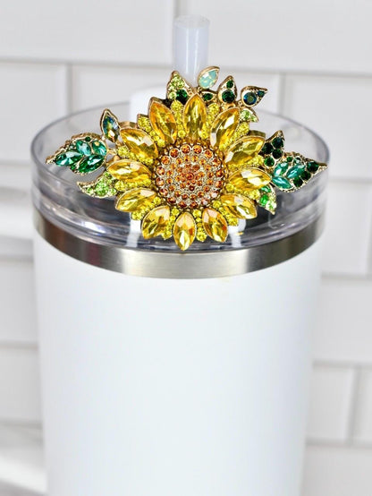 A white tumbler with a decorative sunflower lid. The sunflower is made of yellow and orange crystals with green crystal leaves. The tumbler has a clear lid and a white straw.
