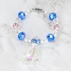 A beaded bracelet featuring blue, pink, and clear crystal beads, with a silver magnetic clasp. A clear, iridescent glass slipper charm hangs from the bracelet. The bracelet is on a white marble surface.