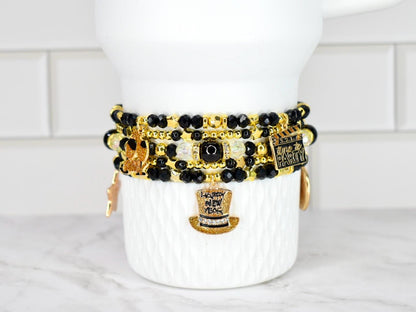 Stack of beaded bracelets in black, gold, and clear beads with charms. Charms include a top hat, a film clapper, and a star. The bracelets are displayed on a white ceramic container.