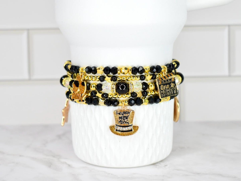 Stack of beaded bracelets in black, gold, and clear beads with charms. Charms include a top hat, a film clapper, and a star. The bracelets are displayed on a white ceramic container.
