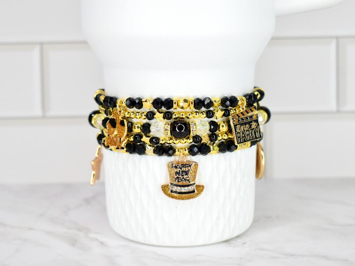 Stack of beaded bracelets in black, gold, and clear beads with charms. Charms include a top hat, a film clapper, and a star. The bracelets are displayed on a white ceramic container.