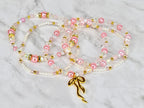 Pink and white beaded necklaces with gold accents and a gold bow pendant. The necklaces feature a mix of pearls, clear beads, and pink beads. The jewelry is displayed on a marble surface.