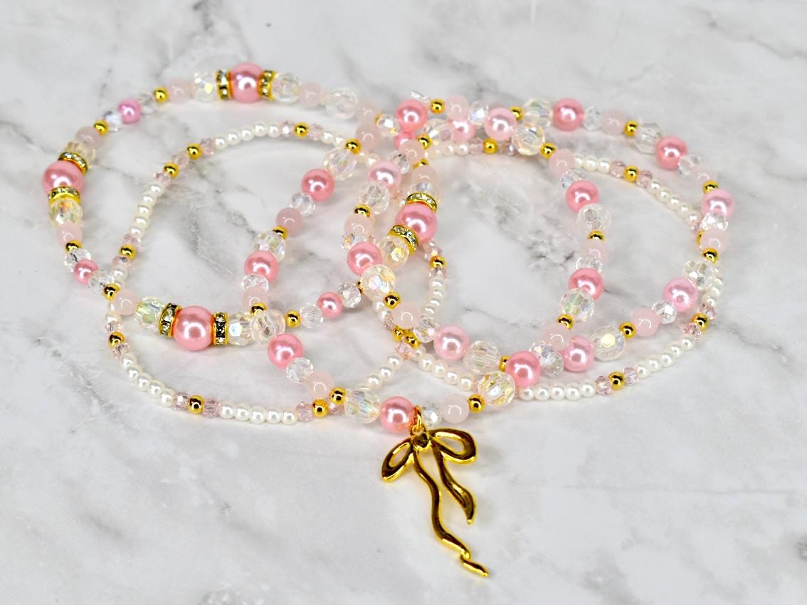 Pink and white beaded necklaces with gold accents and a gold bow pendant. The necklaces feature a mix of pearls, clear beads, and pink beads. The jewelry is displayed on a marble surface.