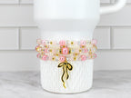 A stack of four beaded bracelets in shades of pink, white, and clear, accented with gold-colored beads and a gold bow charm. The bracelets are displayed on a white, textured surface.