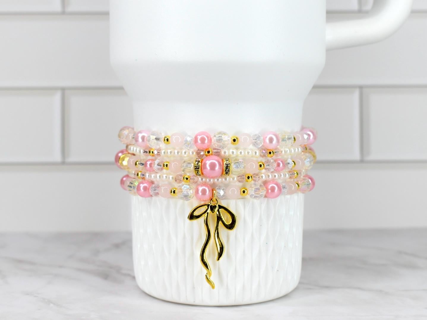 A stack of four beaded bracelets in shades of pink, white, and clear, accented with gold-colored beads and a gold bow charm. The bracelets are displayed on a white, textured surface.