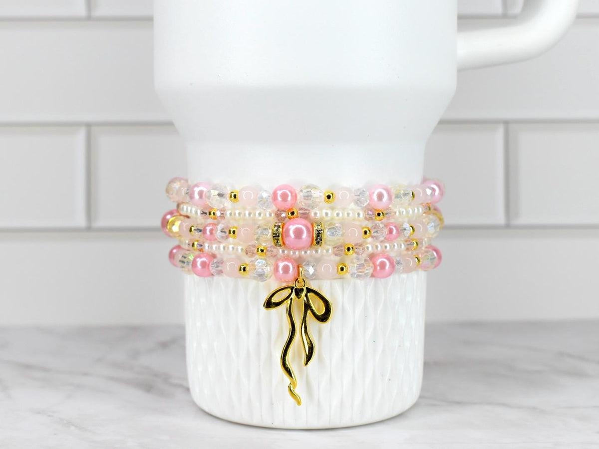 A stack of four beaded bracelets in shades of pink, white, and clear, accented with gold-colored beads and a gold bow charm. The bracelets are displayed on a white, textured surface.