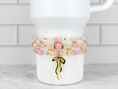 A stack of four beaded bracelets in shades of pink, white, and clear, accented with gold-colored beads and a gold bow charm. The bracelets are displayed on a white, textured surface.