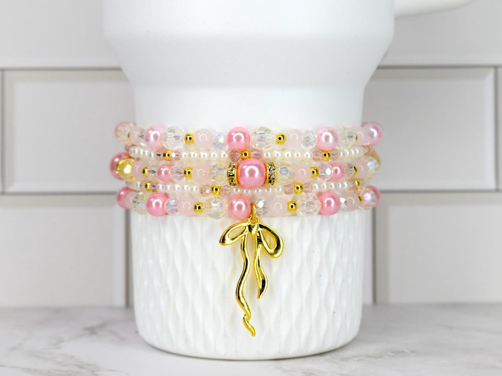 A stack of beaded bracelets in shades of pink, white, and gold. The bracelets are adorned with pearls, clear beads, and gold accents. A gold bow charm hangs from the bottom bracelet. The bracelets are displayed on a white textured surface.