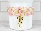 Stack of four beaded bracelets in shades of pink, white, and gold. The bracelets feature a mix of pearls, clear beads, and gold accents. A gold-colored charm in the shape of a ribbon hangs from the bottom bracelet.