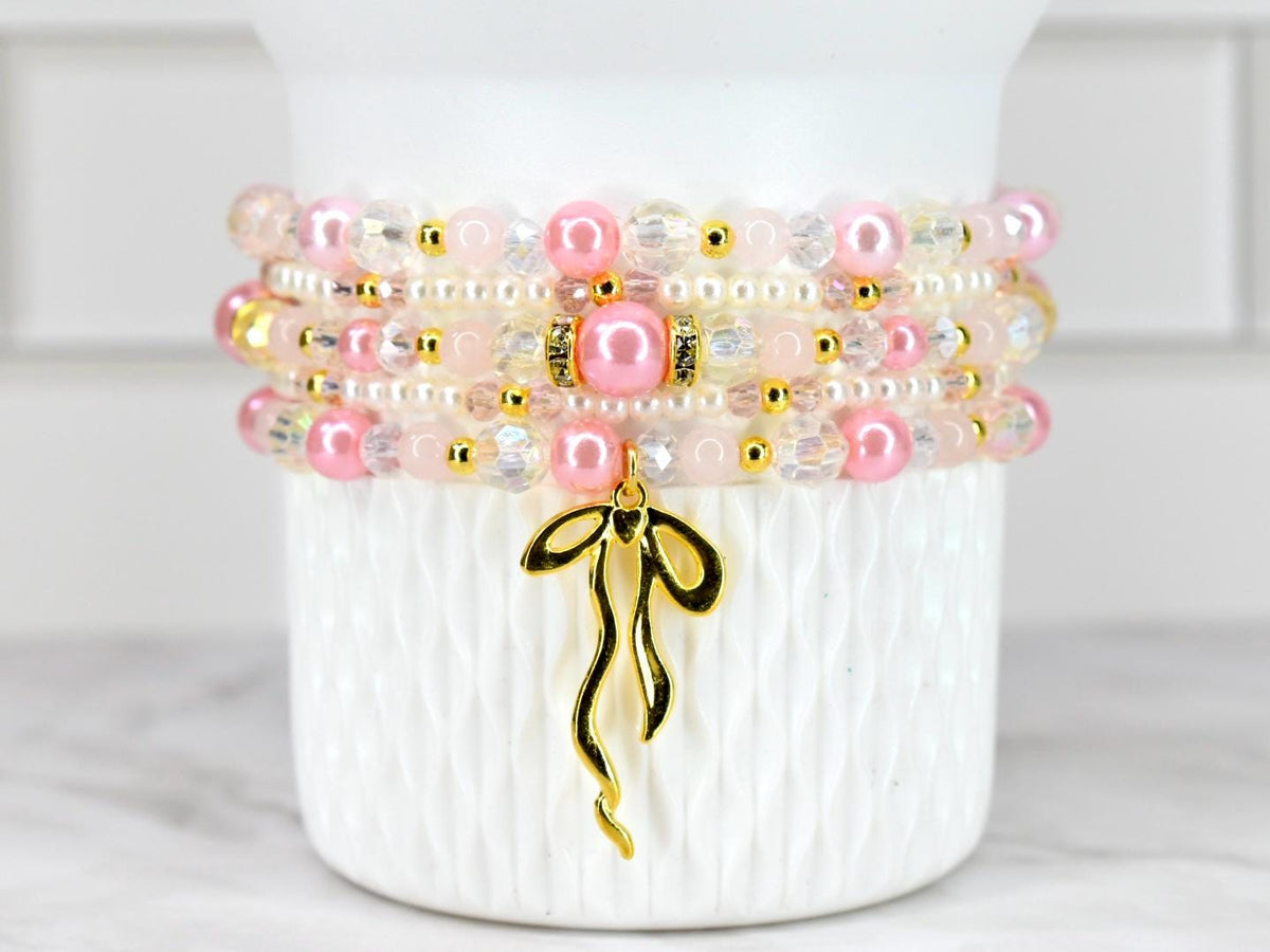 Stack of four beaded bracelets in shades of pink, white, and gold. The bracelets feature a mix of pearls, clear beads, and gold accents. A gold-colored charm in the shape of a ribbon hangs from the bottom bracelet.