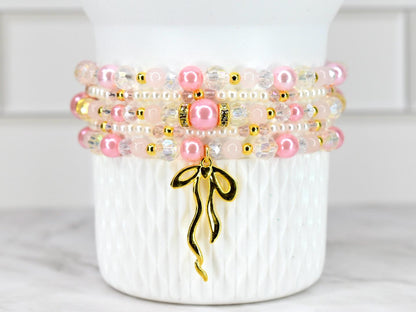 Stack of four beaded bracelets in shades of pink, white, and gold. The bracelets feature a mix of pearls, clear beads, and gold accents. A gold-colored charm in the shape of a ribbon hangs from the bottom bracelet.
