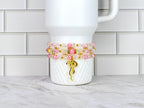 A stack of beaded bracelets in shades of pink, clear, and gold, adorned with a gold charm. The bracelets are wrapped around a white tumbler with a textured pattern.