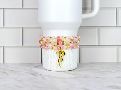 A stack of beaded bracelets in shades of pink, clear, and gold, adorned with a gold charm. The bracelets are wrapped around a white tumbler with a textured pattern.