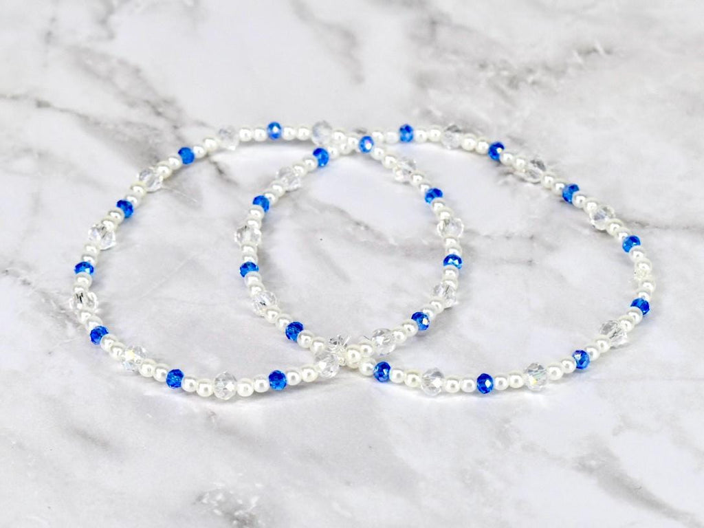 Two beaded bracelets. Each bracelet features white pearls, clear crystal beads, and vibrant blue beads. The bracelets are displayed on a white and gray marble surface.