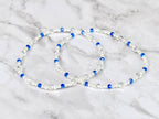 Two beaded bracelets. Each bracelet features white pearls, clear crystal beads, and vibrant blue beads. The bracelets are displayed on a white and gray marble surface.