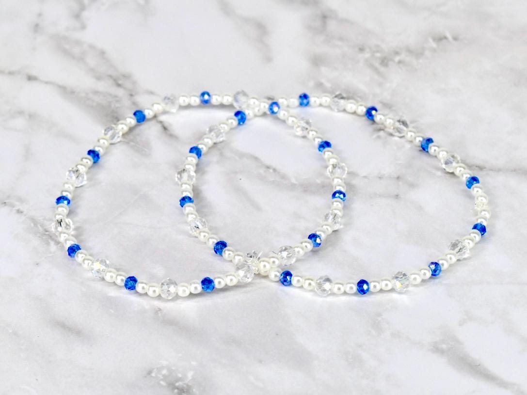 Two beaded bracelets. Each bracelet features white pearls, clear crystal beads, and vibrant blue beads. The bracelets are displayed on a white and gray marble surface.
