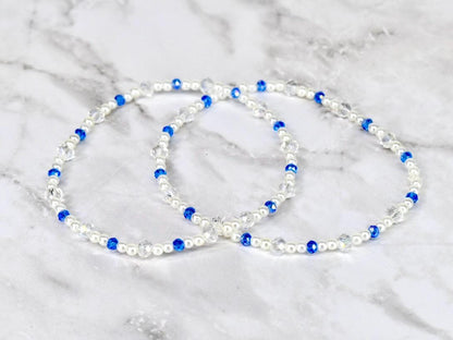 Two beaded bracelets. Each bracelet features white pearls, clear crystal beads, and vibrant blue beads. The bracelets are displayed on a white and gray marble surface.