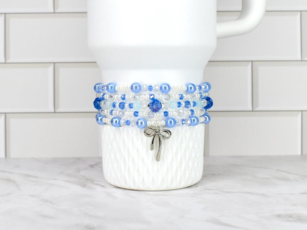 A white tumbler adorned with a beaded bracelet featuring shades of blue and clear beads. A silver bow charm dangles from the bracelet. The tumbler sits on a white and gray surface.